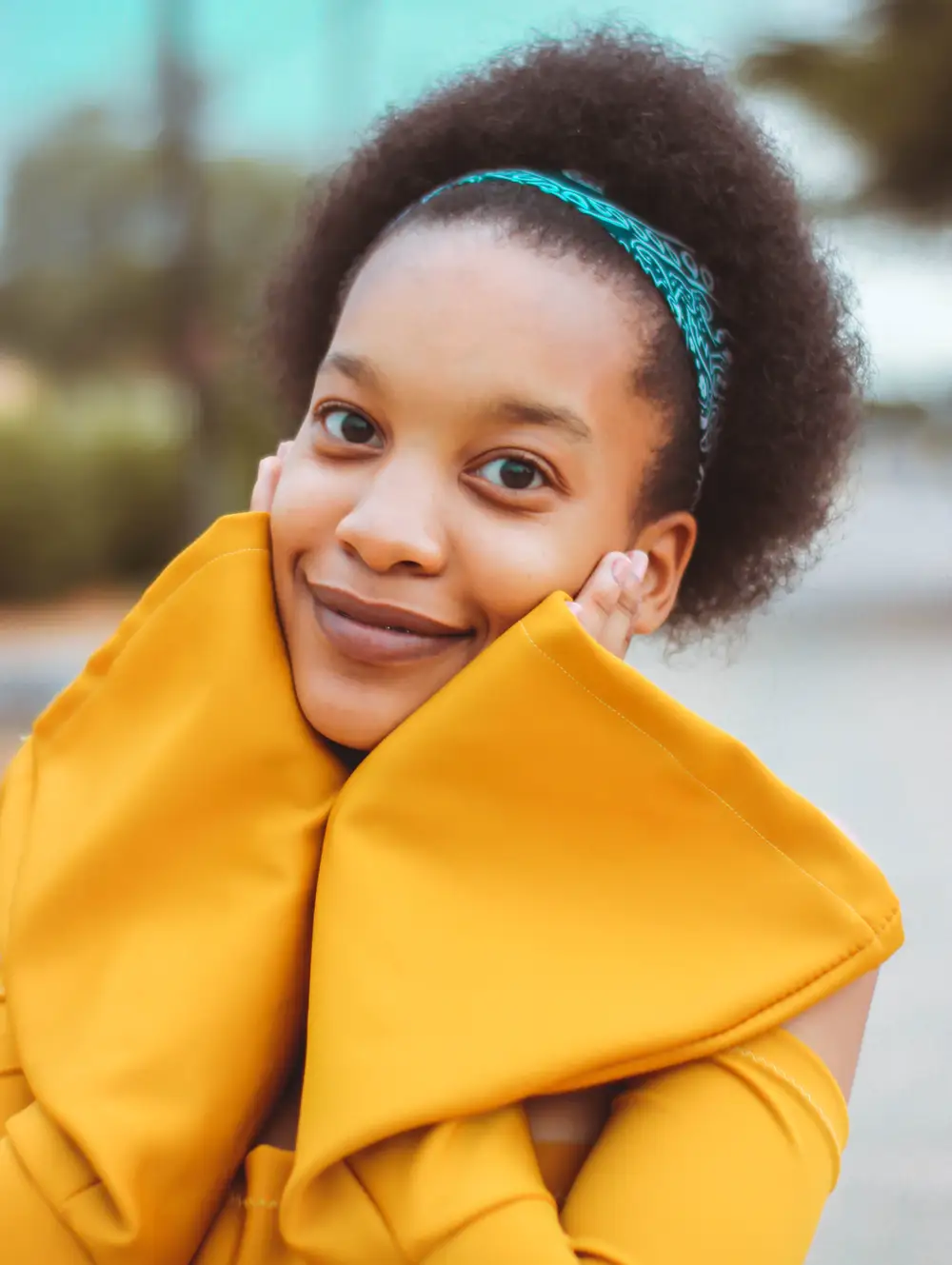 Pretty beige-skin girl with afro hair held by bandana wearing a yellow  dress and also placing hands on her cheek while giving a half-smile