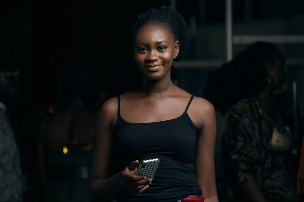Black lady on black top with red belt smiling at the camera and holding an iPhone