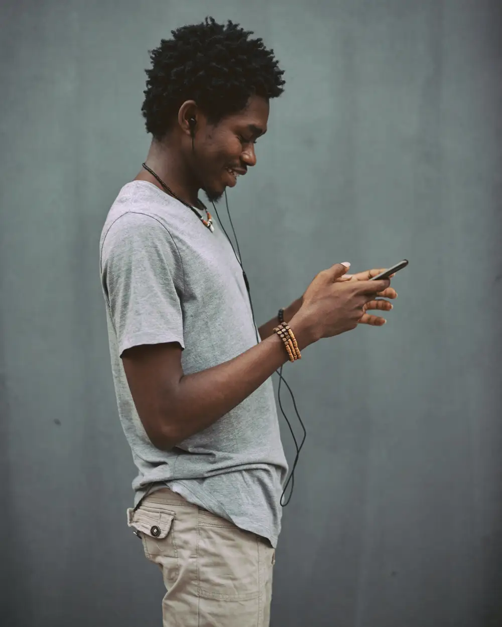 Black man standing against a grey wall holding his earphones and phone.